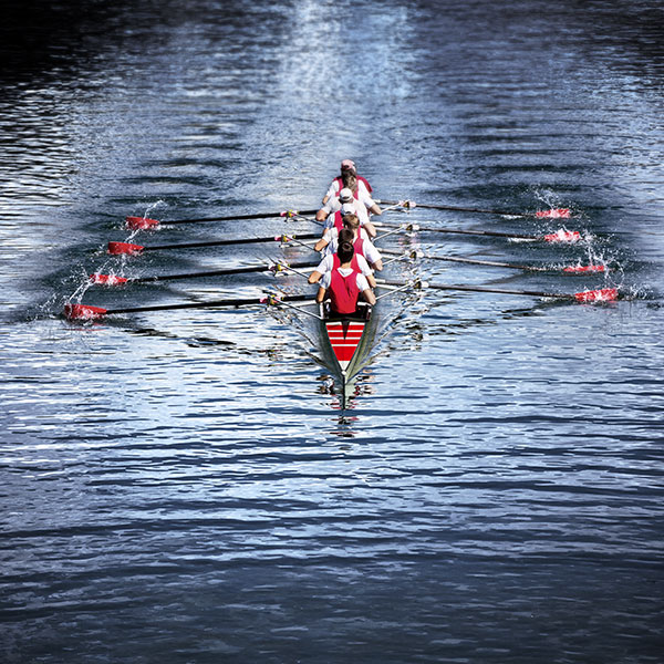 Ruderboot mit acht Athleten in rot-weißer Sportkleidung und synchronen RuderBewegungen mit Wasserspritzern, Vogelperspektive