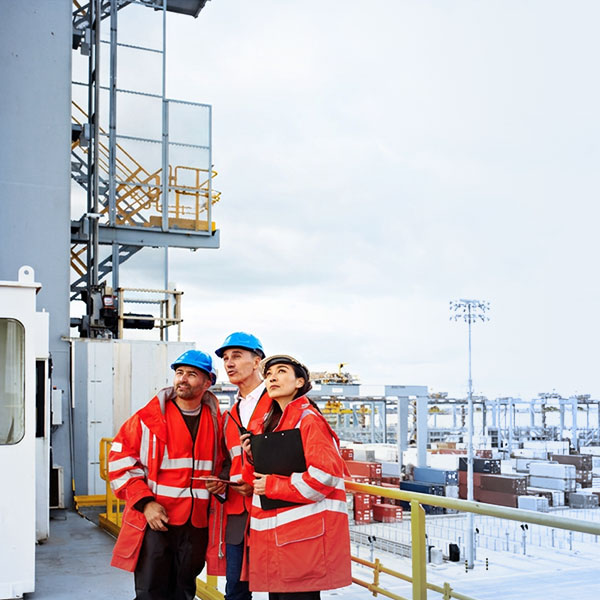 Drei Hafen-Inspekteure in vollständiger Schutzausrüstung (rote Westen, blaue Helme) beaufsichtigen Containerladeprozess an modernem Terminal
