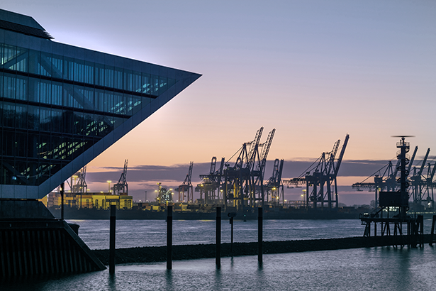 Modernes Gebäude aus Glas vorne links, dahinter Hafenbereich mit Pfählen und Wasser. Hafenkräne als Silhouetten im Dämmerungslicht am Horizont.